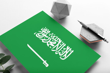 Saudi Arabia flag on minimalist paper background. National invitation letter with stylish pen on stone. Communication concept.