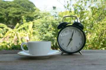 A white cup and a vintage alarm clock on wooden background on a bright day with a nature background.