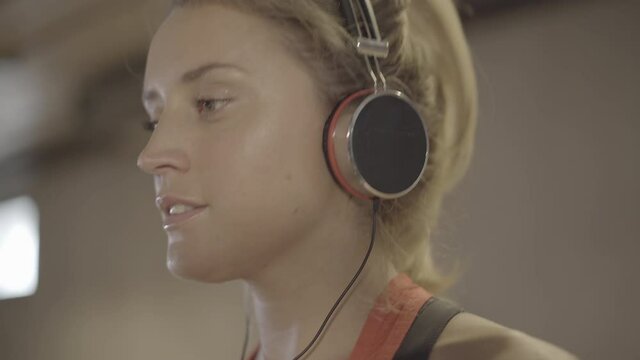 Extreme close-up of young blond woman in headphones training in gym and looking at camera. Portrait of confident Caucasian charming sportswoman exercising on treadmill. Sport, beauty.