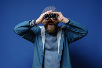 Bearded man looks through binoculars, standing on an isolated blue background, with a surprised...