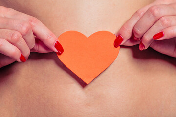 Nice woman holds a paper heart of red at her flat belly. The first signs of skin stretching from pregnancy can be seen on her smooth stomach.