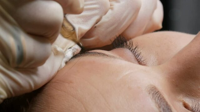 Beautician in black gloves makes permanent makeup correction of the shape of the eyebrows to a young beautiful girl. Microblading, eyebrow tattoo in a beauty salon.