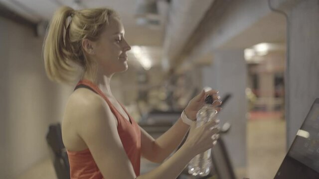 Sportive young Caucasian woman drinking water as walking on treadmill in gym. Side view of beautiful blond sportswoman having break in training. Exercising, sport, lifestyle, endurance.