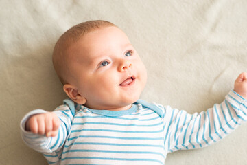 Happy, amused baby boy in blue striped bodysuit lying down on grey blanket, smiling to someone , something on right side. Three months old, blue-eyed caucasian baby with brown hair.