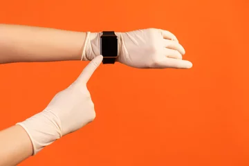 Foto op Plexiglas Profile side view closeup of human hand in white surgical gloves holding and showing wirst smart watch and pointing at empty screen. indoor, studio shot, isolated on orange background. © khosrork