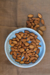Dried almonds in a pale blue ceramic plate.  A bunch of almonds scattered on a pale brown canvas. Copy space. Vertical image.