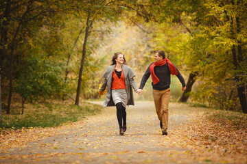 Couple weared in autumn-style clothes (oranfe scarf and vest) running through the autumn landscape. Alley covered with yellow foliage. Autumn walk outdoors. Two lovers in autumn park. Romantic dating