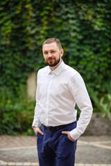 Young man weared in classic suit on background of the City garden. Fashion style guy happy groom in white shirt. Street style businessman portrait. Modern urban wedding walk in old town. Wall greenery