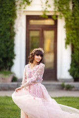 Fototapeta premium Beautiful woman in flying airy pink dress on background of door overgrown with wild grapes. Pretty young caucasian girl plays with her dress. Girl dance in flowing dress