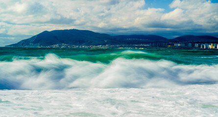 Storm waves at high speed are washed out in white foam. In the background, mountains, multi-storey buildings of the city and port of Novorossiysk.