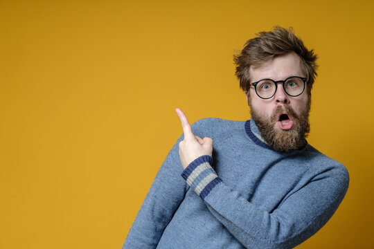 Odd bearded man in glasses points with an index finger to an empty place and stands in a bizarre pose. Copy space.