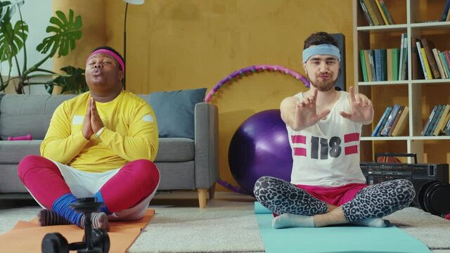 Comical male couple of funny friends jerks wearing retro style sportswear, sitting in lotus pose and practicing relaxing meditation.