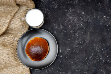 A homemade sweet bun on a black plate, a glass of milk and a burlap cloth on a black texture background. Flat lay, studio shot, copy space. Minimalistic organic breakfast concept.