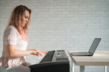 A focused woman plays the electronic piano and watches an online course on her laptop. Distance learning music quarantined. Stay home. A lesson in playing a keyboard instrument.