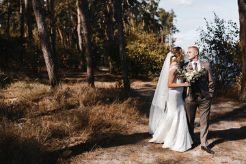 Bride in the wedding dress and veil and groom in checkered suit walking in the pine forest. Bride and groom portrait