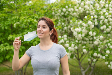 A woman in a gray T-shirt happily takes off her medical mask at the end of quarantine. A girl celebrates the end of quarantine while walking in a park. Free breathing without allergies.