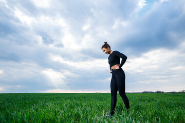 Young attractive brunette woman standing in nature, wearing black leggings and a black top. Summer day, green grass. Athletic body and healthy lifestyle.
