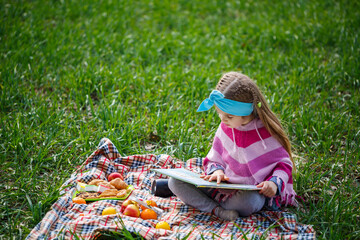 little girl child sits on a bedspread and reads a book with a fairy tale, green grass in the field, sunny spring weather, smile and joy of the child, blue sky with clouds