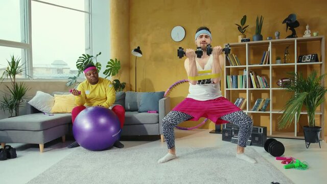 Vigorous active bearded man lifting up heavy dumbbells and stretching legs with bands. His lazy funny friend fat afro-american man resting on couch almost falling asleep.