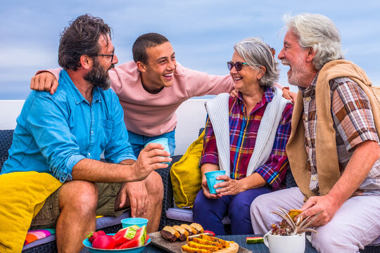 group of four people of all ages together at home in the balcony laughng and having fun and eating food like fruit and cookies - teenager enjoying with seniors and middle age man