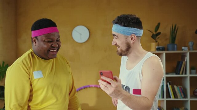 Handsome funny multi-race friends wearing retro sports outfits and laughing of fun content on smartphone browsing social media. Fun, fitness, technology.