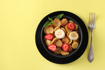 Trendy tiny pancakes for breakfast with strawberry and banana in dark bowl on yellow background. View from above. Space for text