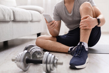Power training. Guy with fitness tracker and smartphone sits on floor with dumbbells