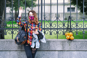 Young woman wearing a plaid handmade face mask and colorful shirt with mesh bag full of oranges and a backpack standing on the street.
