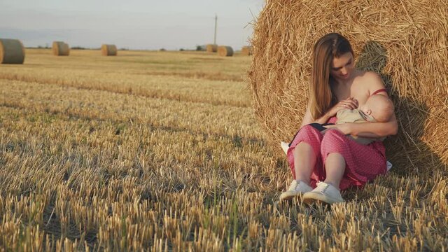 Portrait of pretty young mother feeds baby son with breast at haystack in field