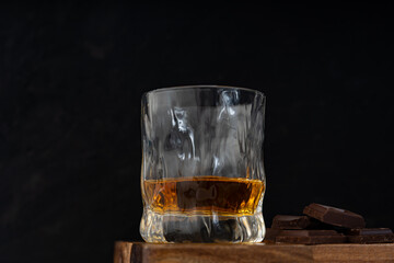 A glass of rum together with dark chocolate slices are laying on wooden bar counter against dark...