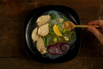 Black plate of Vietnamese Pho soup with chicken on a wooden table with a spoon.