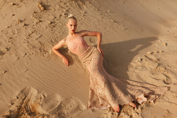 Blonde woman in splendid gold dress posing in desert, lying on the sand at the sunset.