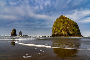 Fototapeta na wymiar Haystack Rock and the Needles reflected in the wet sand, with wave foam and blue sky and clouds, Canon Beach, Oregon