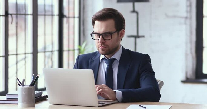 Concentrated young ceo manager employee in suit working on laptop, thinking of hard problem solution. 30s male executive looking at computer screen, doing online financial market research in office.