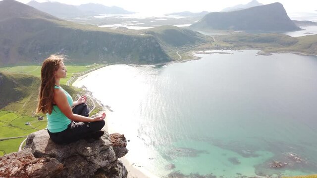 A young, white female hiker is sitting down and meditating on a rock on the edge of a cliff on top of Mannen mountain, overlooking Haukland beach during a warm and sunny summer day in Lofoten, Norway.