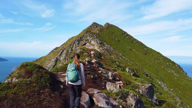 A young brunette woman is walking with her hiking boots and backpack on a trail leading up to the top of mountain Mannen. On the edge of the cliff leading down to Haukland Beach in Lofoten, Norway.
