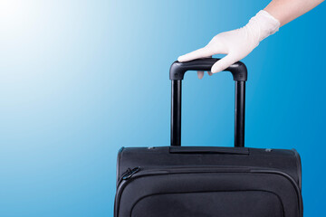 Hand with white latex gloves holding travel suitcase  on light blue background. Journey and viral prevention during pandemic.