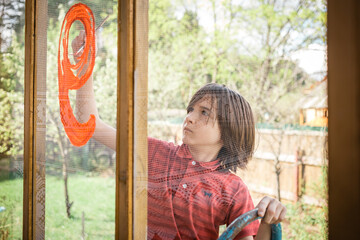 A boy in a red T-shirt draws with colorful gouache on the window