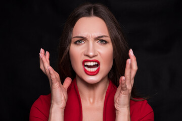Emotional head shot portrait of a brunette caucasian woman in red dress and with red lips on black background. She make angry, mad, cry emotion