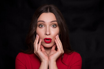 Emotional head shot portrait of a brunette caucasian woman in red dress and with red lips on black background. She make surprise, astonishment emotions with hands near her face