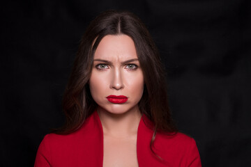 Emotional head shot portrait of a brunette caucasian woman in red dress and with red lips on black background. She make sad emotion