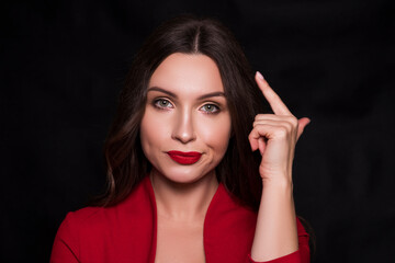 Emotional head shot portrait of a brunette caucasian woman in red dress and with red lips on black background. She make disdain, arrogant emotions with finger near her temple