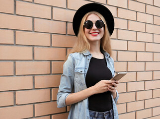 Portrait of smiling young woman with phone looking at camera wearing a black round hat, jeans jacket over brick wall background