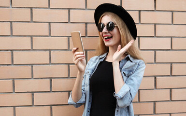 Portrait of surprised young woman with phone on city street wearing a black round hat, jeans jacket over brick wall background