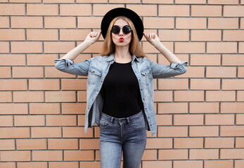 Fototapeta na wymiar Stylish young woman blowing red lips sending sweet air kiss wearing a black round hat, jeans jacket, female model on brick wall background