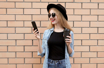Portrait of smiling young woman with phone and coffee cup wearing a black round hat, jeans jacket over brick wall background
