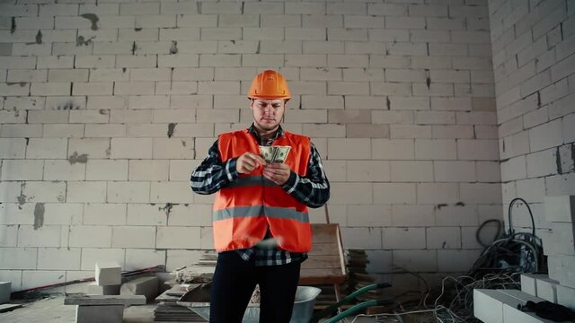 A male builder is standing in a building amid a construction site and is slowly scattering money.