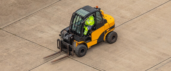 Worker dressed in light green safety clothing driving a yellow fork lift on flat concrete surface. Aerial top view.