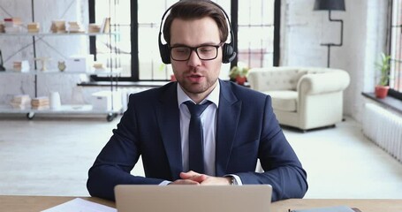 Focused serious skilled young salesman in formal suit wearing wireless earphones with microphone, advertising professional services to business clients by video conference call in modern office.