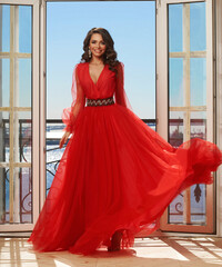 Young beautiful stunning girl in long red dress posing in interior with open door and view on sunny blue sky. Woman in elegant ball gown